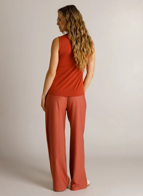 A woman with long wavy blonde hair is shown from the back wearing a sleeveless rust-orange top and matching wide-leg pants against a plain light gray studio background.